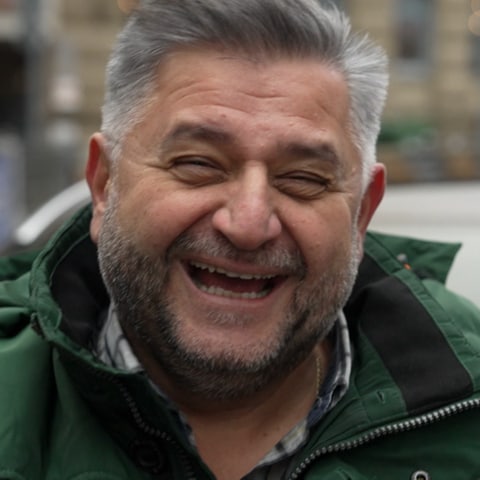 Iraklis ist Taxifahrer in Stuttgart. Er steht in der Stadt und lächelt herzlich in die Kamera. 