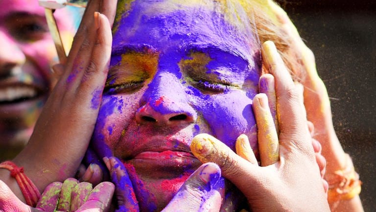28. März: Das Leuchten des Frühlings! Eine junge Frau schließt die Augen und feiert auf dem Holi Festival, dem Fest der Farben. Ihr Gesicht - umzingelt von Händen - ist bedeckt mit Neonpulver in lilanen, pinken und gelben Tönen. 