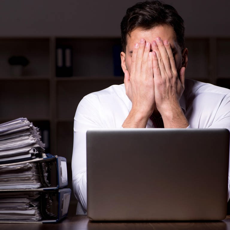 Arbeitssucht: Ein Mann sitzt nachts an seinem Schreibtisch vor einem Notebook und einem Stapel Aktenordnern | Arbeitssucht, Arbeit und Gesundheit